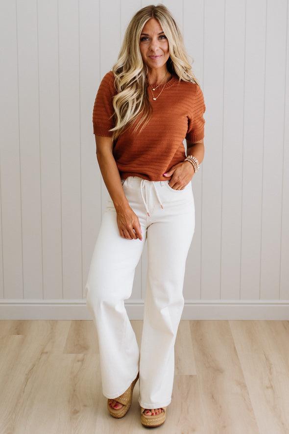 Woman wearing a rust-colored top and white pants standing against a light wooden paneled wall.