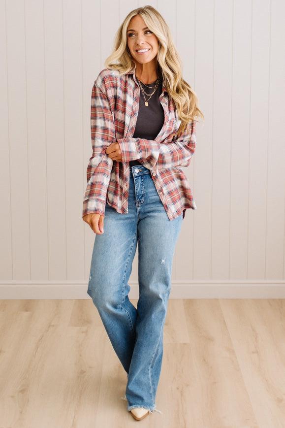 A woman modeling a red and white plaid button-down shirt over a dark top and blue jeans, standing in a room with light wood floors and white paneled walls.