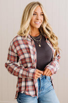 A woman wearing a red, white, and blue plaid button-up flannel shirt over a dark gray top and blue jeans, standing against a light beige paneled wall.