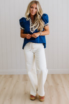 Woman wearing a blue top with lace details and white pants against a light wooden floor and wall background.
