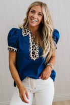 Woman wearing a blue blouse with leopard print details and white pants against a neutral background