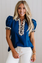 Woman wearing a blue blouse with leopard print details and white pants against a light background