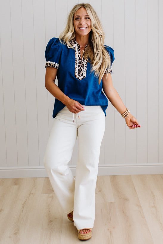 Woman wearing a blue top with lace details and white pants against a light wooden floor and wall background.