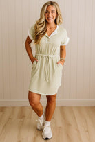 Woman wearing a light green dress with a white shirt underneath, standing against a beige wall.