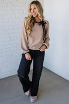 Woman wearing a beige sweater with black ribbons and dark jeans against a white brick wall.