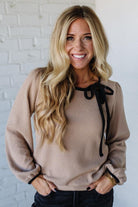 Woman wearing a beige sweater with black ribbons against a white brick wall