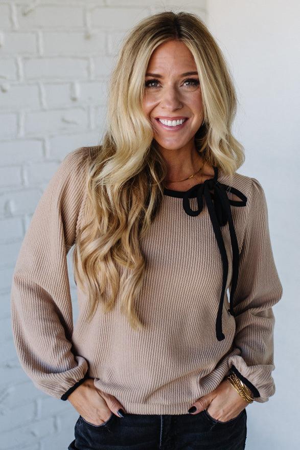 Woman wearing a beige sweater with black ribbons against a white brick wall