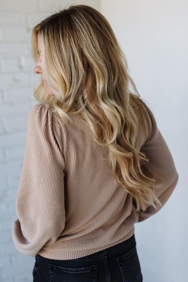 Woman wearing a beige ribbed sweater against a white brick wall