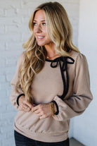 Woman wearing a beige sweater with black ribbons against a white brick wall.