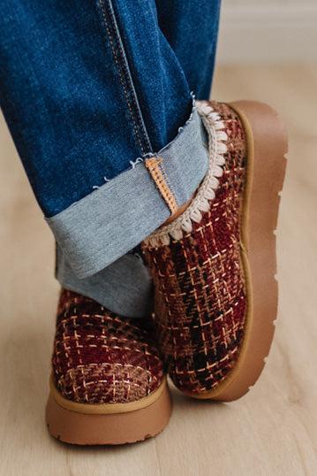 A close-up of a person wearing cuffed blue jeans and chunky knit slip-on shoes in a burgundy, brown, and beige tweed pattern with tan soles, standing on a light wood floor.