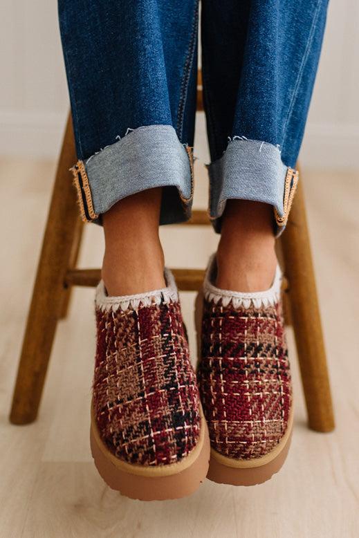A person wearing cuffed blue jeans and slip-on platform slippers with a red, burgundy, and black tweed pattern, sitting on a wooden stool.