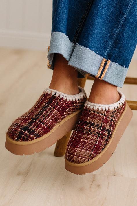 A person wearing clog-style slip-on shoes with a burgundy, black, and tan tweed pattern, white fuzzy trim, and tan platform soles, paired with rolled-up blue jeans on a light wood floor.
