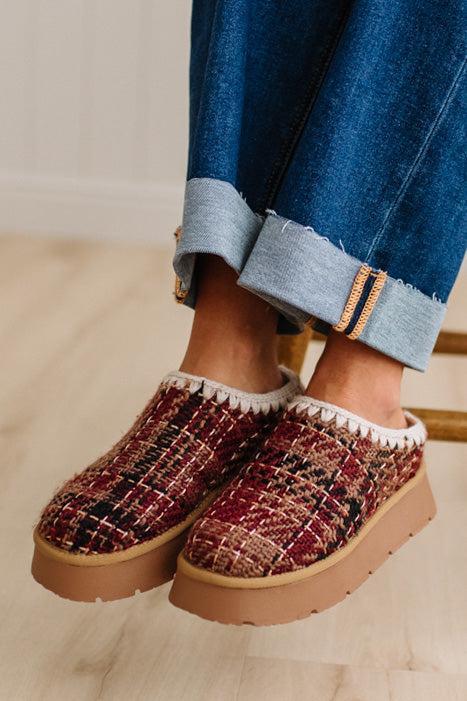 A person wearing clog-style slip-on shoes with a burgundy, black, and tan tweed pattern, white fuzzy trim, and tan platform soles, paired with rolled-up blue jeans on a light wood floor.