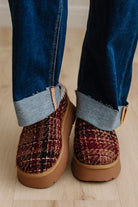 A close-up shot of a person's feet wearing cuffed blue jeans and brown woven slippers with tan soles, standing on a light wood floor.