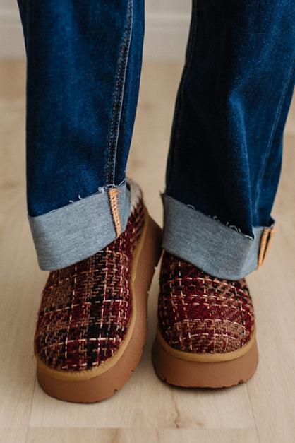 A close-up shot of a person's feet wearing cuffed blue jeans and brown woven slippers with tan soles, standing on a light wood floor.