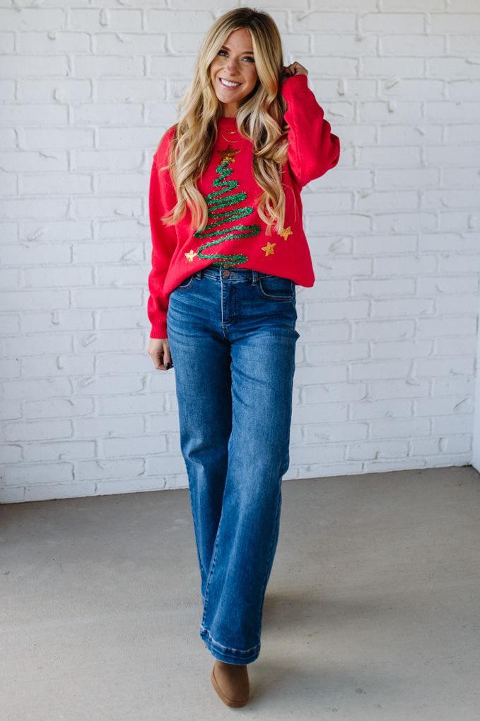 Woman wearing a red sweater with a Christmas tree design and blue jeans against a white brick wall.