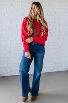 Woman wearing a red sweater with a Christmas tree design and blue jeans against a white brick wall.