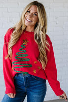 Woman wearing a red sweater with Christmas tree design against a white brick wall.