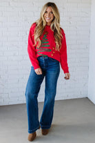 Woman wearing a red sweater with a Christmas tree design and blue jeans against a white brick wall.
