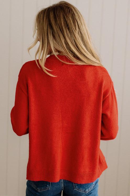 Blonde woman in dark jeans and a bold red-orange cardigan with black button toggles down with front and side pockets.