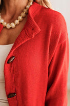 Blonde woman in dark jeans and a bold red-orange cardigan with black button toggles down with front and side pockets.