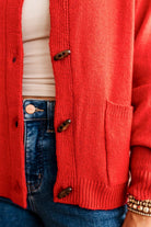 Blonde woman in dark jeans and a bold red-orange cardigan with black button toggles down with front and side pockets.