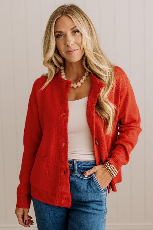 Blonde woman in dark jeans and a bold red-orange cardigan with black button toggles down with front and side pockets.