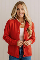 Blonde woman in dark jeans and a bold red-orange cardigan with black button toggles down with front and side pockets.