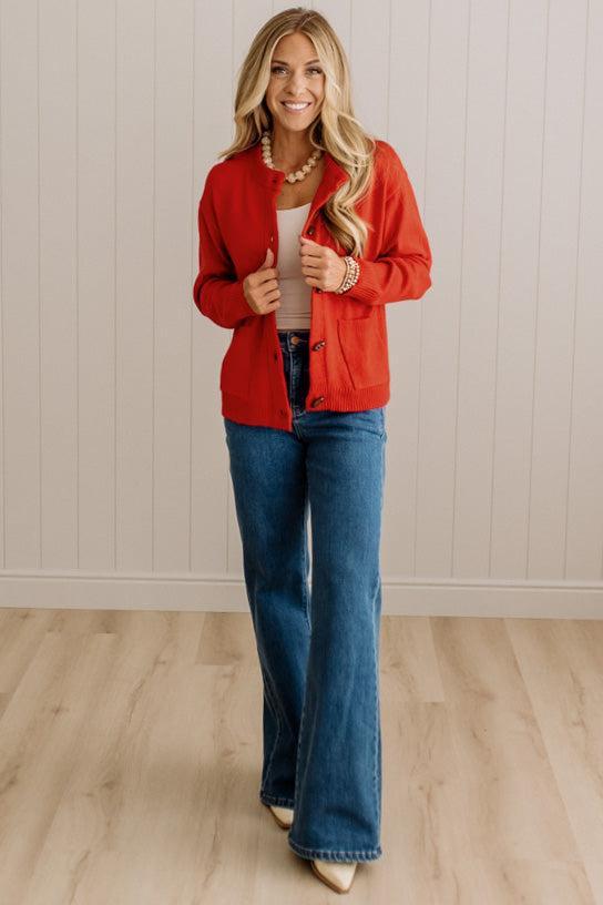 Blonde woman in dark jeans and a bold red-orange cardigan with black button toggles down with front and side pockets.