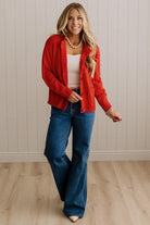 Blonde woman in dark jeans and a bold red-orange cardigan with black button toggles down with front and side pockets.