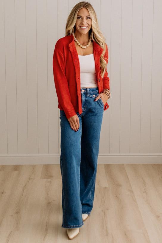 Blonde woman in dark jeans and a bold red-orange cardigan with black button toggles down with front and side pockets.