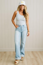 Woman wearing a white tank top, light blue jeans, and a white cap with a yellow logo against a wooden paneled wall.