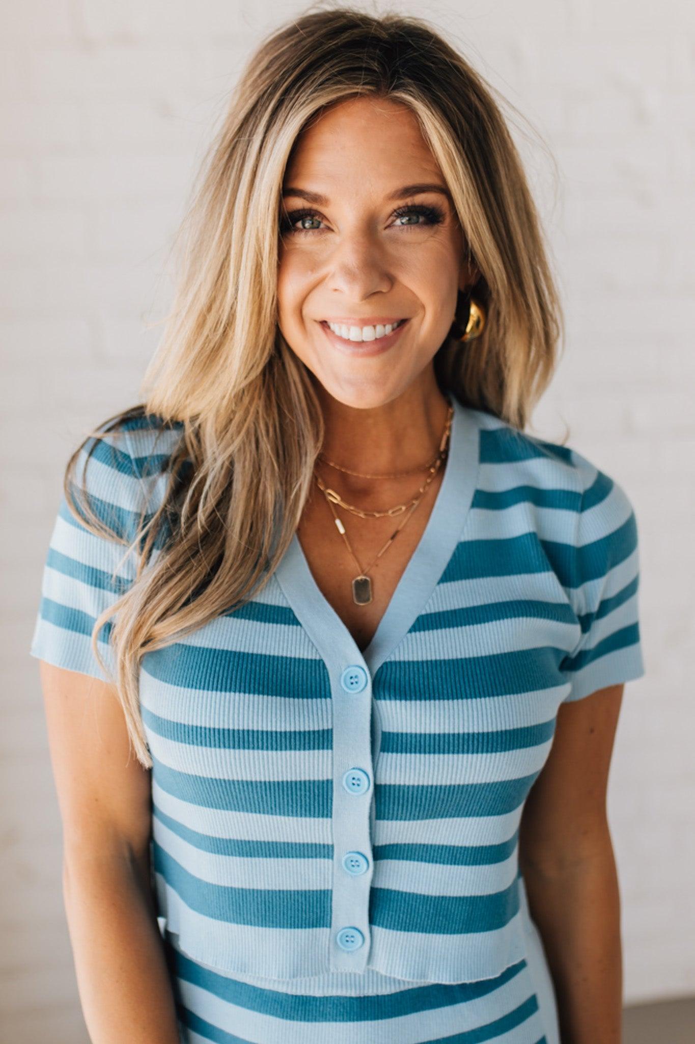 Blonde woman wearing a light blue and medium blue horizontal striped top with button front sweater and short sleeves.