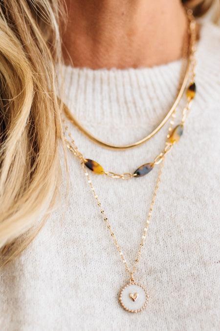 A refined layered necklace featuring a sleek gold herringbone chain paired with a delicate link strand accented by warm tortoiseshell-style acetate beads. A petite heart coin pendant adds a soft feminine touch, creating an elegant balance between modern texture and timeless shine. The adjustable length allows the layers to sit perfectly along the neckline for effortless styling.