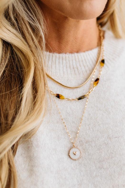 A refined layered necklace featuring a sleek gold herringbone chain paired with a delicate link strand accented by warm tortoiseshell-style acetate beads. A petite heart coin pendant adds a soft feminine touch, creating an elegant balance between modern texture and timeless shine. The adjustable length allows the layers to sit perfectly along the neckline for effortless styling.