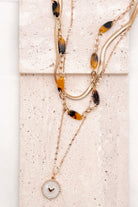 A refined layered necklace featuring a sleek gold herringbone chain paired with a delicate link strand accented by warm tortoiseshell-style acetate beads. A petite heart coin pendant adds a soft feminine touch, creating an elegant balance between modern texture and timeless shine. The adjustable length allows the layers to sit perfectly along the neckline for effortless styling.