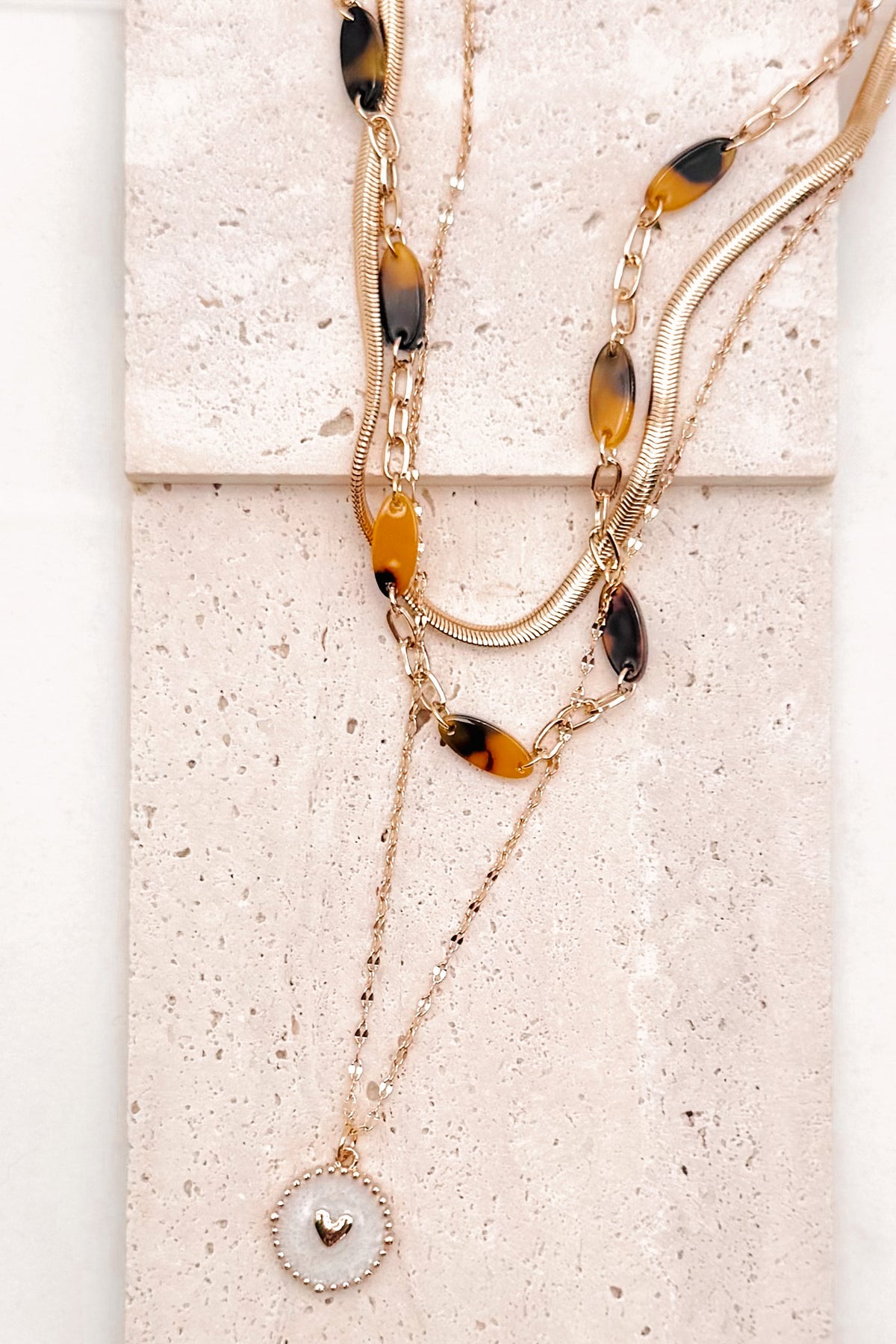 A refined layered necklace featuring a sleek gold herringbone chain paired with a delicate link strand accented by warm tortoiseshell-style acetate beads. A petite heart coin pendant adds a soft feminine touch, creating an elegant balance between modern texture and timeless shine. The adjustable length allows the layers to sit perfectly along the neckline for effortless styling.