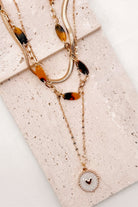 A refined layered necklace featuring a sleek gold herringbone chain paired with a delicate link strand accented by warm tortoiseshell-style acetate beads. A petite heart coin pendant adds a soft feminine touch, creating an elegant balance between modern texture and timeless shine. The adjustable length allows the layers to sit perfectly along the neckline for effortless styling.