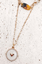 A refined layered necklace featuring a sleek gold herringbone chain paired with a delicate link strand accented by warm tortoiseshell-style acetate beads. A petite heart coin pendant adds a soft feminine touch, creating an elegant balance between modern texture and timeless shine. The adjustable length allows the layers to sit perfectly along the neckline for effortless styling.