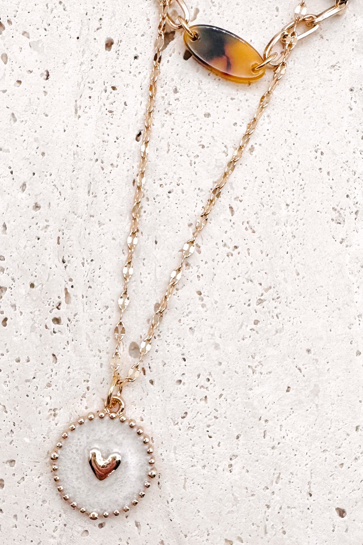 A refined layered necklace featuring a sleek gold herringbone chain paired with a delicate link strand accented by warm tortoiseshell-style acetate beads. A petite heart coin pendant adds a soft feminine touch, creating an elegant balance between modern texture and timeless shine. The adjustable length allows the layers to sit perfectly along the neckline for effortless styling.