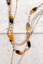 A refined layered necklace featuring a sleek gold herringbone chain paired with a delicate link strand accented by warm tortoiseshell-style acetate beads. A petite heart coin pendant adds a soft feminine touch, creating an elegant balance between modern texture and timeless shine. The adjustable length allows the layers to sit perfectly along the neckline for effortless styling.