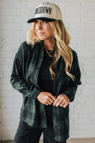 Woman wearing a green plaid shirt, black pants, and a cap with 'Touchdown' text against a white brick wall.
