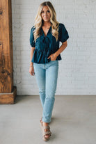 Woman wearing a teal blouse and light blue jeans against a white brick wall.