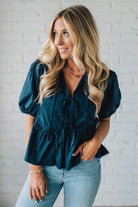 Woman wearing a teal blouse and light blue jeans against a white brick wall.