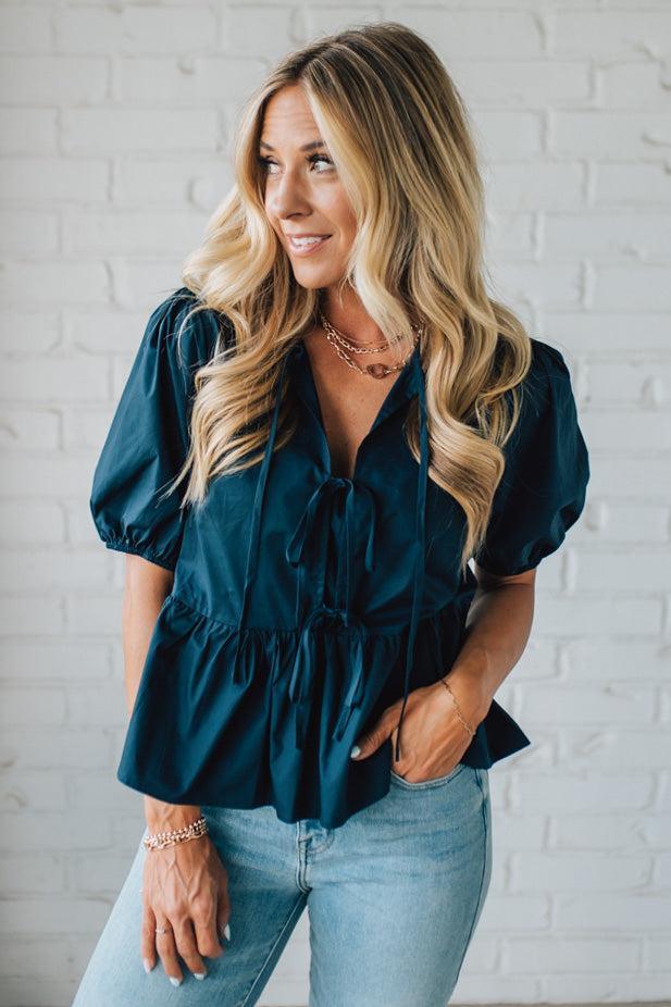 Woman wearing a teal blouse and light blue jeans against a white brick wall.