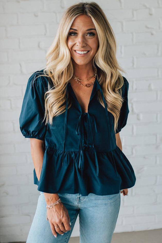 Woman wearing a teal blouse with puff sleeves against a white brick wall.