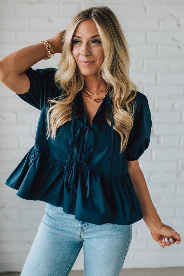 Woman wearing a teal blouse and light blue jeans against a white brick wall.