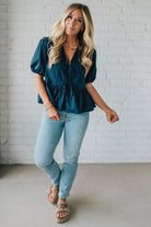 Woman wearing a teal blouse and light blue jeans against a white brick wall.