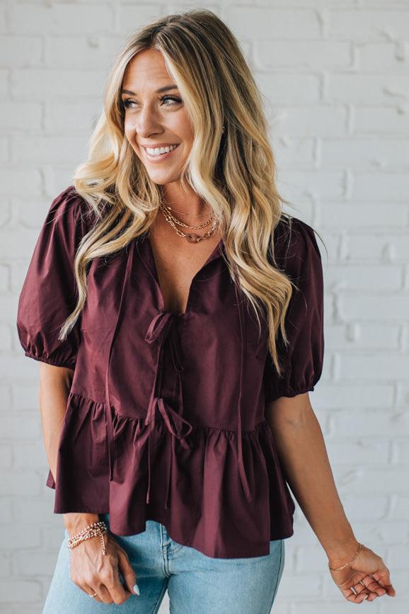 Woman wearing a burgundy blouse with ruffled details against a white brick wall.