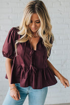 Woman wearing a burgundy blouse with puffed sleeves and light blue jeans against a white brick wall.